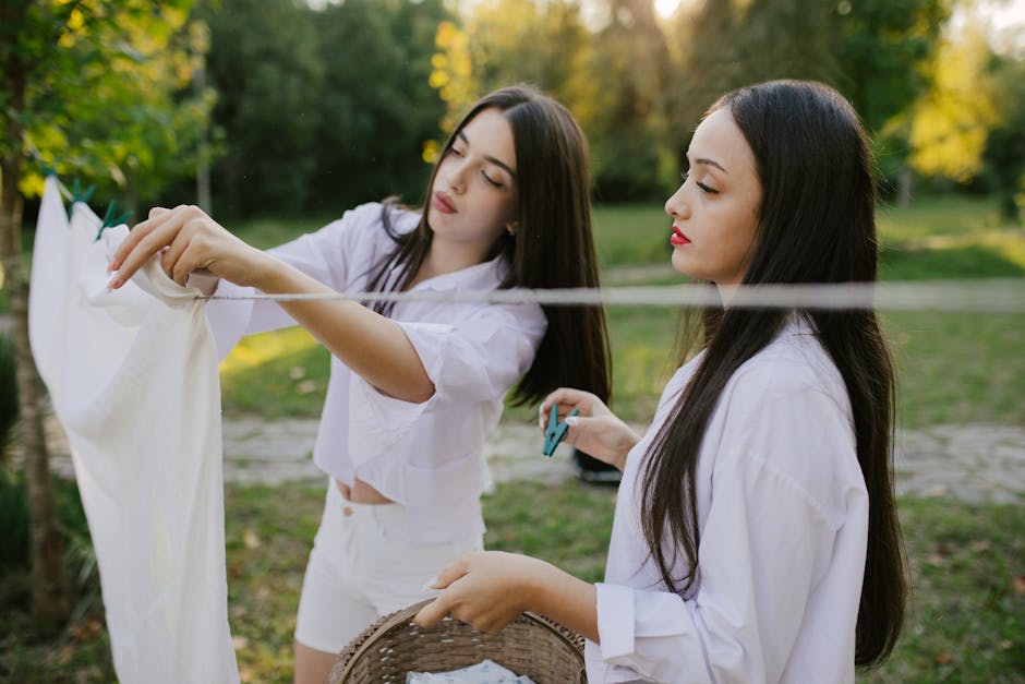 Two young women hanging laundry on a clothesline outdoors in a summer setting, enjoying a sunny day.