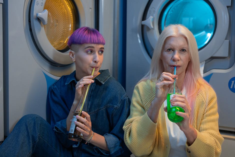 Two women with unique hairstyles enjoying drinks in a laundry room setting, showcasing modern lifestyle.
