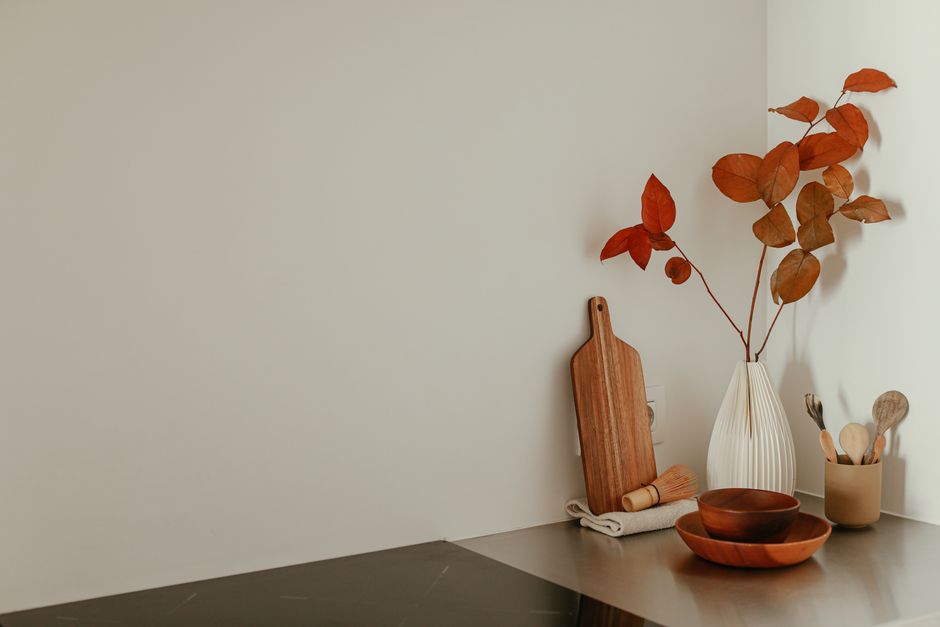 A minimalist kitchen corner featuring red leaves in a ceramic vase, wooden utensils, and a chopping board.
