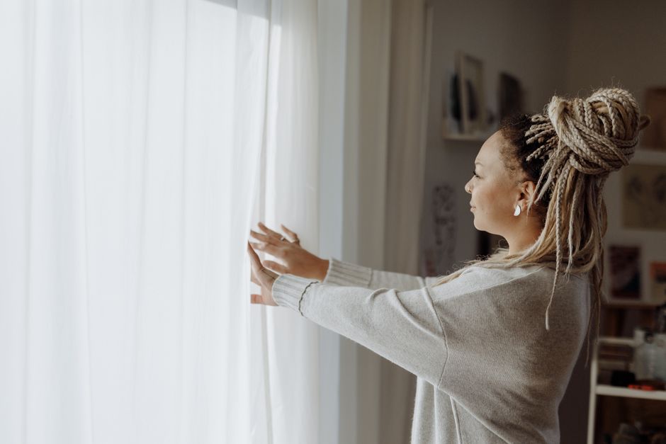 Side view of a woman in loungewear opening curtains to let in morning light.