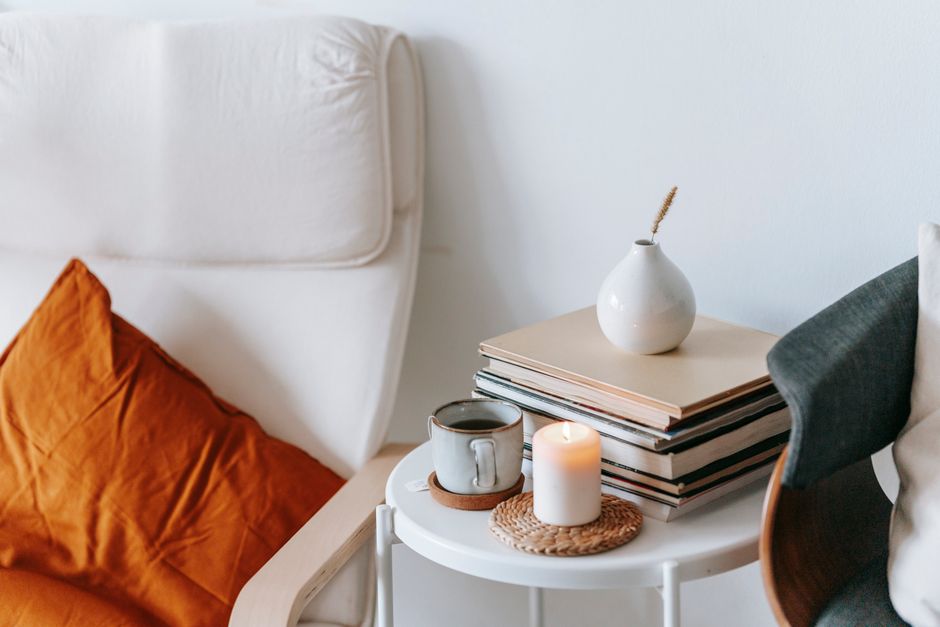Cup of tea and flaming candle against heap of books with dry spike in vase between armchairs at home on white background