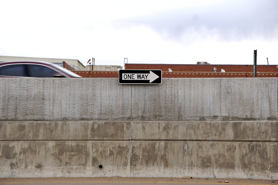 Cityscape featuring a one-way road sign, passing car, and urban concrete wall.