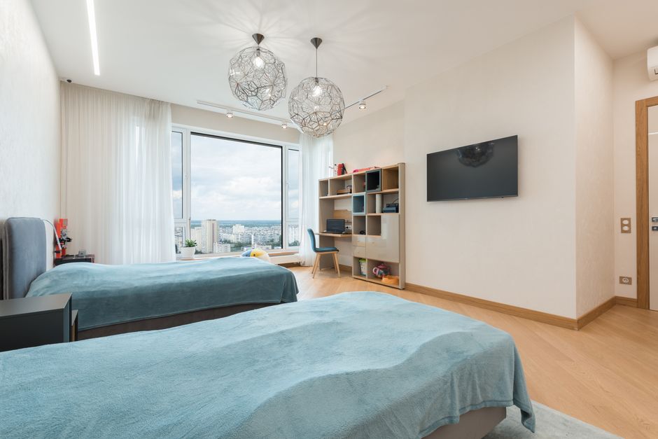 Comfortable twin beds placed in front of TV set and bookshelf in contemporary bedroom with big windows and chandeliers