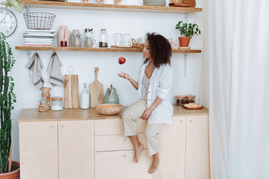 A woman with curly hair juggling an apple in a cozy, minimalist kitchen.