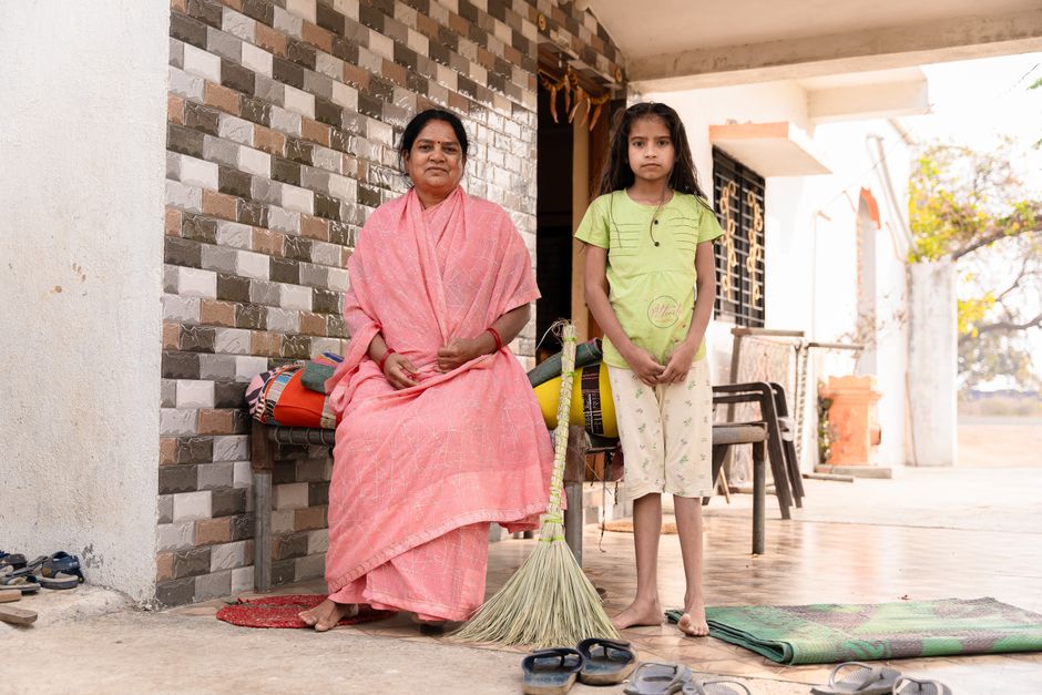 A woman and a girl posing outside their rural Indian home with a broom.