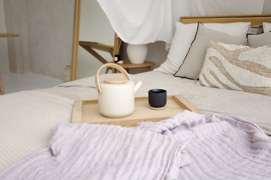 A cozy bedroom setting with a teapot on a tray and soft lavender blankets.